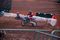 A motocross rider in a red outfit and helmet navigates a dirt track indoors, leaning into a turn. The indoor arena has orange walls with horizontal black lines and a safety barrier surrounds the track. Another rider in a similar outfit is visible in the background, along with a standing person in an orange vest.