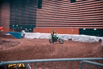 A motorcyclist wearing a helmet and protective gear rides a dirt bike on an indoor motocross track. The track is surrounded by barriers and situated in a large hall with dark horizontal slatted walls. The lighting gives the scene an energetic and focused atmosphere.