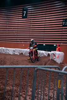 A motocross rider wearing protective gear is navigating a dirt track inside an indoor facility. The number 7 is prominently displayed on a large wooden panel wall in the background. Another person in a red outfit stands near the track, possibly a marshal or staff member. Barriers and fabric line the edge of the track for safety.