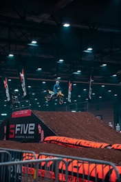A motocross rider is mid-air on a dirt bike, performing a jump inside an indoor arena. The arena is dimly lit with overhead lights, and there are dirt ramps and barriers visible. Several flags with logos are placed around the area, and there are people in the background watching the event.