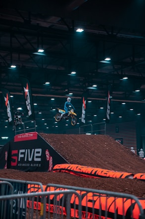 A motocross rider is mid-air on a dirt bike, performing a jump inside an indoor arena. The arena is dimly lit with overhead lights, and there are dirt ramps and barriers visible. Several flags with logos are placed around the area, and there are people in the background watching the event.