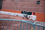 A motorcyclist is navigating a dirt track indoors, leaning into a turn. The surroundings include a protective barrier wrapped in white fabric and a person standing in a high-visibility vest observing the event. The walls are adorned with horizontal slats, and there is an overhead display showing numbers.