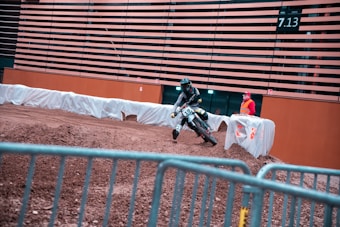 A motorcyclist is navigating a dirt track indoors, leaning into a turn. The surroundings include a protective barrier wrapped in white fabric and a person standing in a high-visibility vest observing the event. The walls are adorned with horizontal slats, and there is an overhead display showing numbers.