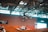 A motorcyclist performs an aerial trick on a dirt bike inside an arena. The rider is airborne, showcasing skill and control over the bike, with the backdrop of industrial ceiling beams and a striped wall displaying advertisements. Spectators wearing helmets and protective gear observe from below, near a mound of dirt.