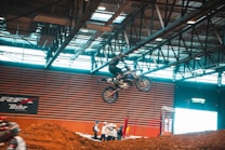 A motorcyclist performs an aerial trick on a dirt bike inside an arena. The rider is airborne, showcasing skill and control over the bike, with the backdrop of industrial ceiling beams and a striped wall displaying advertisements. Spectators wearing helmets and protective gear observe from below, near a mound of dirt.