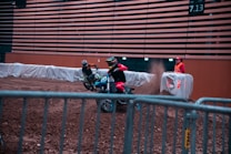 Two motorcyclists in racing gear are riding dirt bikes inside an indoor arena. They are maneuvering around a dirt track behind safety barriers. A race official in a high-visibility vest stands nearby, watching the action, while dirt is kicked up behind the bikes.