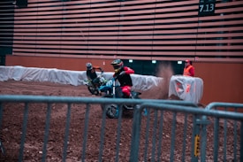 Two motorcyclists in racing gear are riding dirt bikes inside an indoor arena. They are maneuvering around a dirt track behind safety barriers. A race official in a high-visibility vest stands nearby, watching the action, while dirt is kicked up behind the bikes.