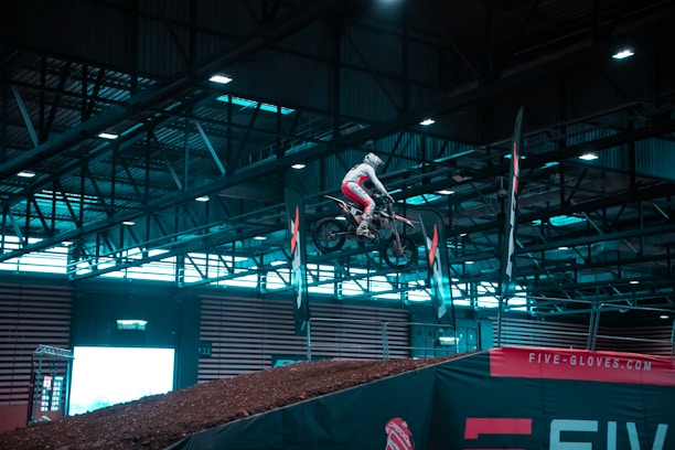 A motorcyclist is seen mid-air performing a jump inside a large indoor arena. The rider is dressed in white and red gear, riding a dirt bike over a dirt ramp. The background features tall metal structures and beams, indicative of an industrial setting, with banners and advertisements including one for Five-Gloves.com.