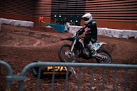 A motorcyclist wearing a helmet and protective gear rides a dirt bike on an indoor dirt track. Barriers line the track, and a person in an orange jacket stands in the background. The environment is dimly lit, and the track surface is rough with various mounds and ridges.