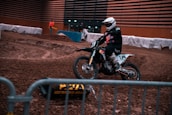 A motorcyclist wearing a helmet and protective gear rides a dirt bike on an indoor dirt track. Barriers line the track, and a person in an orange jacket stands in the background. The environment is dimly lit, and the track surface is rough with various mounds and ridges.