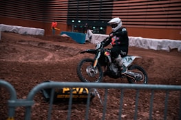 A motorcyclist wearing a helmet and protective gear rides a dirt bike on an indoor dirt track. Barriers line the track, and a person in an orange jacket stands in the background. The environment is dimly lit, and the track surface is rough with various mounds and ridges.
