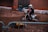 A motorcyclist wearing a helmet and protective gear rides a dirt bike on an indoor dirt track. Barriers line the track, and a person in an orange jacket stands in the background. The environment is dimly lit, and the track surface is rough with various mounds and ridges.