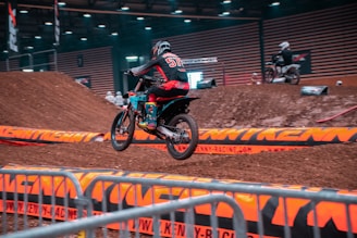 Motocross riders jumping over dirt ramps in a rugged outdoor track.