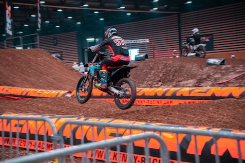 Motocross riders jumping over dirt ramps in a rugged outdoor track.