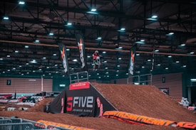 A motocross rider is performing a high-flying stunt on a dirt ramp inside a large indoor arena. The area is well-lit, highlighting banners and branding for a competition. The event appears to be organized with safety barriers and spectators in the background.