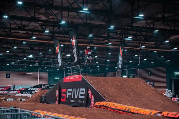 A motocross rider is performing a high-flying stunt on a dirt ramp inside a large indoor arena. The area is well-lit, highlighting banners and branding for a competition. The event appears to be organized with safety barriers and spectators in the background.