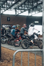 A group of motocross riders in racing gear and helmets are lined up on dirt bikes, preparing to start a race indoors. The setting appears to be a dedicated arena with metal railings and overhead lighting visible.