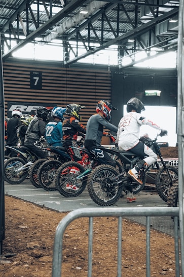 A group of motocross riders in racing gear and helmets are lined up on dirt bikes, preparing to start a race indoors. The setting appears to be a dedicated arena with metal railings and overhead lighting visible.