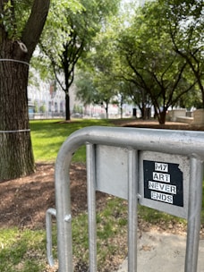 A metal barricade in the foreground features a black and white sticker saying 'MY ART NEVER ENDS.' Behind it, a park with lush green grass and numerous trees provides a serene backdrop. Sunlight filters through the leaves, casting dappled shadows on the ground.