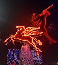 A neon-lit red Pegasus with wings, prominently displayed against a nighttime backdrop. The vibrant sign appears to be mounted on a structure, with a blue-lit building in the background. The Pegasus's shape is dynamically captured, giving a sense of motion.
