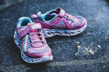 A pair of children's athletic shoes resting on a textured, dark pavement. The shoes are predominantly pink with turquoise interiors, featuring colored laces and thick white soles with heart-shaped cut-outs.