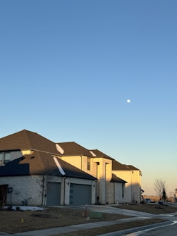 A suburban neighborhood with a row of modern, two-story brick houses. The exterior walls are light-colored with dark gray roofs, and the street is lined with a sidewalk and newly planted trees. The sky is clear with a visible moon, indicating early evening or morning.