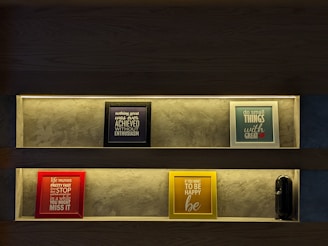 Two shelves with illuminated frames displaying motivational quotes are mounted against a textured wall. The top shelf features a black-framed piece reading 'nothing great was ever achieved without enthusiasm' and a blue-framed piece with 'do small things with great love'. The bottom shelf contains a red-framed quote 'life moves pretty fast if you don't stop and look around once in a while you might miss it' and a yellow-framed quote 'if you want to be happy be'. A small black speaker is also present on the bottom shelf.