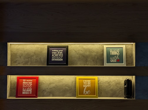Two shelves with illuminated frames displaying motivational quotes are mounted against a textured wall. The top shelf features a black-framed piece reading 'nothing great was ever achieved without enthusiasm' and a blue-framed piece with 'do small things with great love'. The bottom shelf contains a red-framed quote 'life moves pretty fast if you don't stop and look around once in a while you might miss it' and a yellow-framed quote 'if you want to be happy be'. A small black speaker is also present on the bottom shelf.
