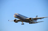 A large commercial airplane is flying through a clear blue sky with its landing gear extended. The aircraft is prominently displayed, showcasing its engines, wings, and fuselage.