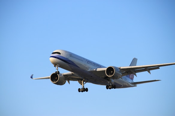 A large commercial airplane is flying through a clear blue sky with its landing gear extended. The aircraft is prominently displayed, showcasing its engines, wings, and fuselage.