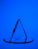 A transparent triangular prism is placed on a circular glass base against a solid blue background. The prism casts sharp reflections and shadows, creating a geometric aesthetic.