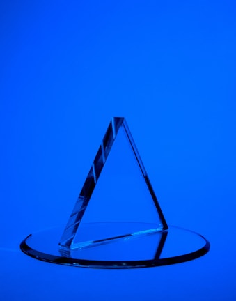 A transparent triangular prism is placed on a circular glass base against a solid blue background. The prism casts sharp reflections and shadows, creating a geometric aesthetic.