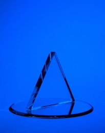 A transparent triangular prism is placed on a circular glass base against a solid blue background. The prism casts sharp reflections and shadows, creating a geometric aesthetic.