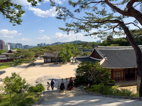 Traditional Korean architecture with tiled roofs set amidst green trees and surrounded by modern buildings in the distance. A small group of people in traditional attire walks down stone steps, under the canopy of tree branches.