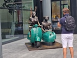 A sculpture featuring two anthropomorphic figures, a rabbit and a dog wearing suits, seated on a vintage motorcycle with a sidecar. The rabbit is holding the handlebars while the dog sits in the sidecar, holding a cup. A person with a backpack is capturing the scene using a smartphone in front of a modern building with reflective glass walls.