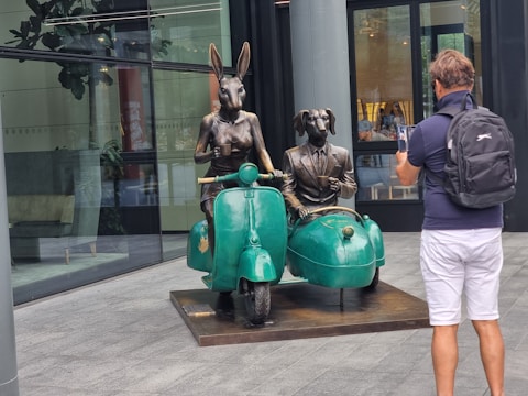 A sculpture featuring two anthropomorphic figures, a rabbit and a dog wearing suits, seated on a vintage motorcycle with a sidecar. The rabbit is holding the handlebars while the dog sits in the sidecar, holding a cup. A person with a backpack is capturing the scene using a smartphone in front of a modern building with reflective glass walls.