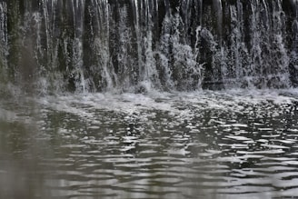A cascading waterfall with multiple streams of water flowing downwards, creating splashes and foam as it hits the surface of the pond below. The water glistens with reflections, and the overall scene conveys a natural and serene environment.