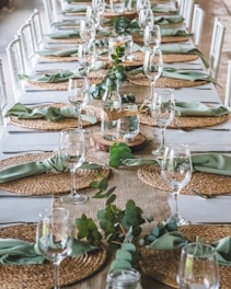 A long dining table elegantly set with woven placemats, green napkins, and clear wine glasses. In the center, there are glass bottles with greenery and small candles, creating a natural, inviting atmosphere. The arrangement suggests preparation for a formal event or gathering.