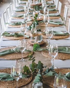 A long dining table elegantly set with woven placemats, green napkins, and clear wine glasses. In the center, there are glass bottles with greenery and small candles, creating a natural, inviting atmosphere. The arrangement suggests preparation for a formal event or gathering.