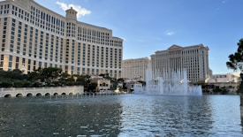 A large, iconic hotel with a curved design stands prominently behind a large, beautifully landscaped lake. Fountains are actively shooting streams of water into the air, creating a dynamic display. Trees and other greenery partially line the foreground, with a clear blue sky overhead.