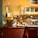 A busy commercial kitchen with chefs in white uniforms preparing food. The scene includes various cooking utensils, appliances, and countertops. There are shelves with decorative vases and a sign overhead. Bright lighting highlights the warm, inviting ambiance of the kitchen.