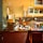 A busy commercial kitchen with chefs in white uniforms preparing food. The scene includes various cooking utensils, appliances, and countertops. There are shelves with decorative vases and a sign overhead. Bright lighting highlights the warm, inviting ambiance of the kitchen.