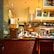 A busy commercial kitchen with chefs in white uniforms preparing food. The scene includes various cooking utensils, appliances, and countertops. There are shelves with decorative vases and a sign overhead. Bright lighting highlights the warm, inviting ambiance of the kitchen.