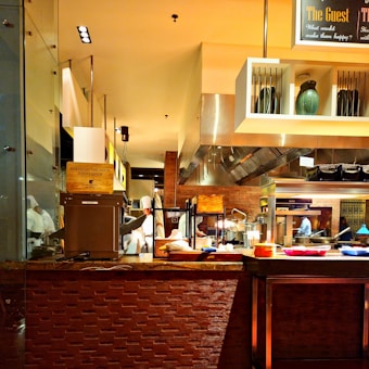 A busy commercial kitchen with chefs in white uniforms preparing food. The scene includes various cooking utensils, appliances, and countertops. There are shelves with decorative vases and a sign overhead. Bright lighting highlights the warm, inviting ambiance of the kitchen.