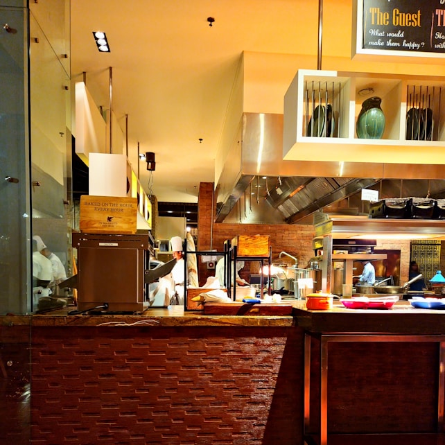 A busy commercial kitchen with chefs in white uniforms preparing food. The scene includes various cooking utensils, appliances, and countertops. There are shelves with decorative vases and a sign overhead. Bright lighting highlights the warm, inviting ambiance of the kitchen.