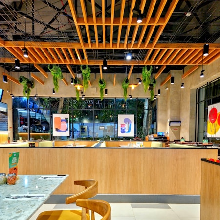 A modern, stylish restaurant interior featuring wooden ceiling designs with hanging green plants and multiple pendant lights. Large windows line the wall, allowing natural light to stream in, complementing the vibrant artworks on the walls. The seating area is neatly arranged with tables and chairs, and there is a warm ambiance created by the combination of wood and greenery.