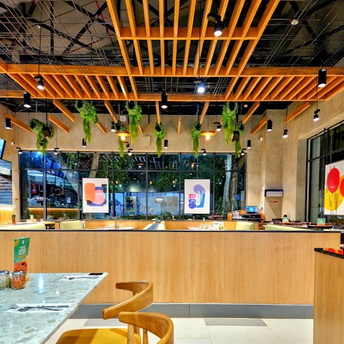 A modern, stylish restaurant interior featuring wooden ceiling designs with hanging green plants and multiple pendant lights. Large windows line the wall, allowing natural light to stream in, complementing the vibrant artworks on the walls. The seating area is neatly arranged with tables and chairs, and there is a warm ambiance created by the combination of wood and greenery.