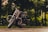 A sleek motorcycle is parked on a wet pavement with lush, green trees in the background. The light reflects off the chrome details, and the scene is serene with a hint of adventure.