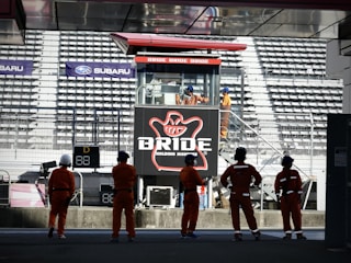 A group of people in orange uniforms and helmets are standing in front of a grandstand. In the background, there is a large advertising billboard with the word 'BRIDE' prominently displayed. The stadium seats are mostly empty, and several individuals are visible on a staircase near the sign.