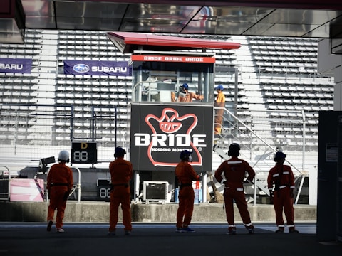 A group of people in orange uniforms and helmets are standing in front of a grandstand. In the background, there is a large advertising billboard with the word 'BRIDE' prominently displayed. The stadium seats are mostly empty, and several individuals are visible on a staircase near the sign.